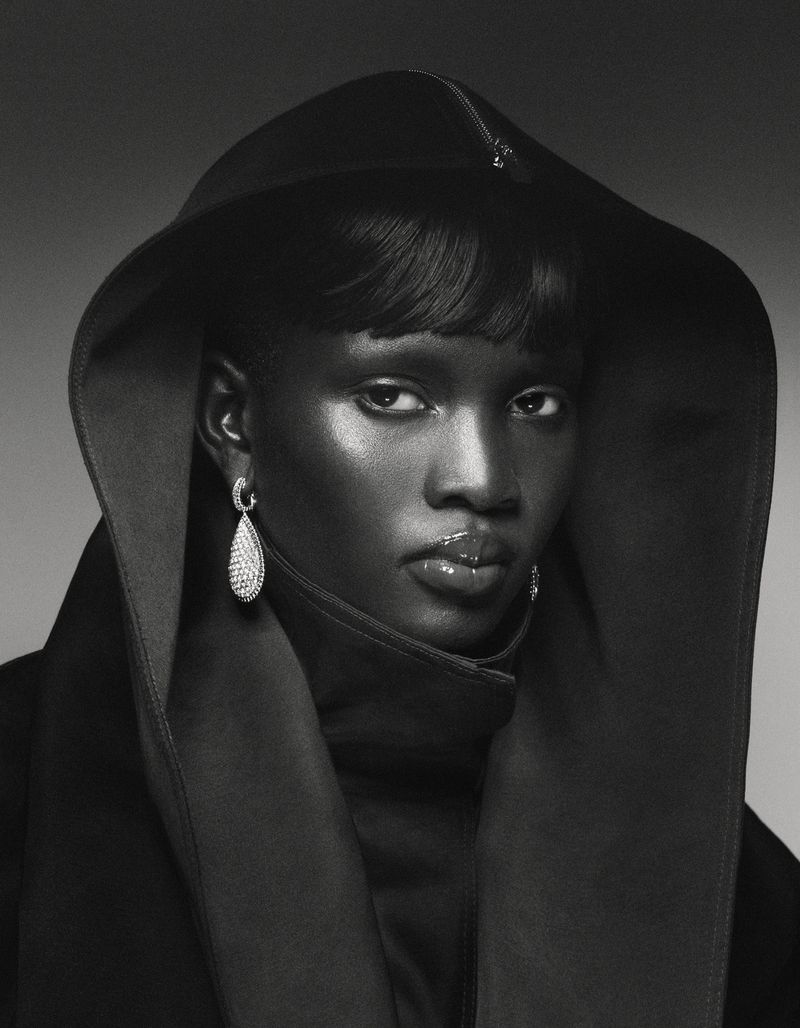 Close-up studio shot of an African-American woman with short black hair, wearing a hood, dark top, and teardrop earrings against a gray background.