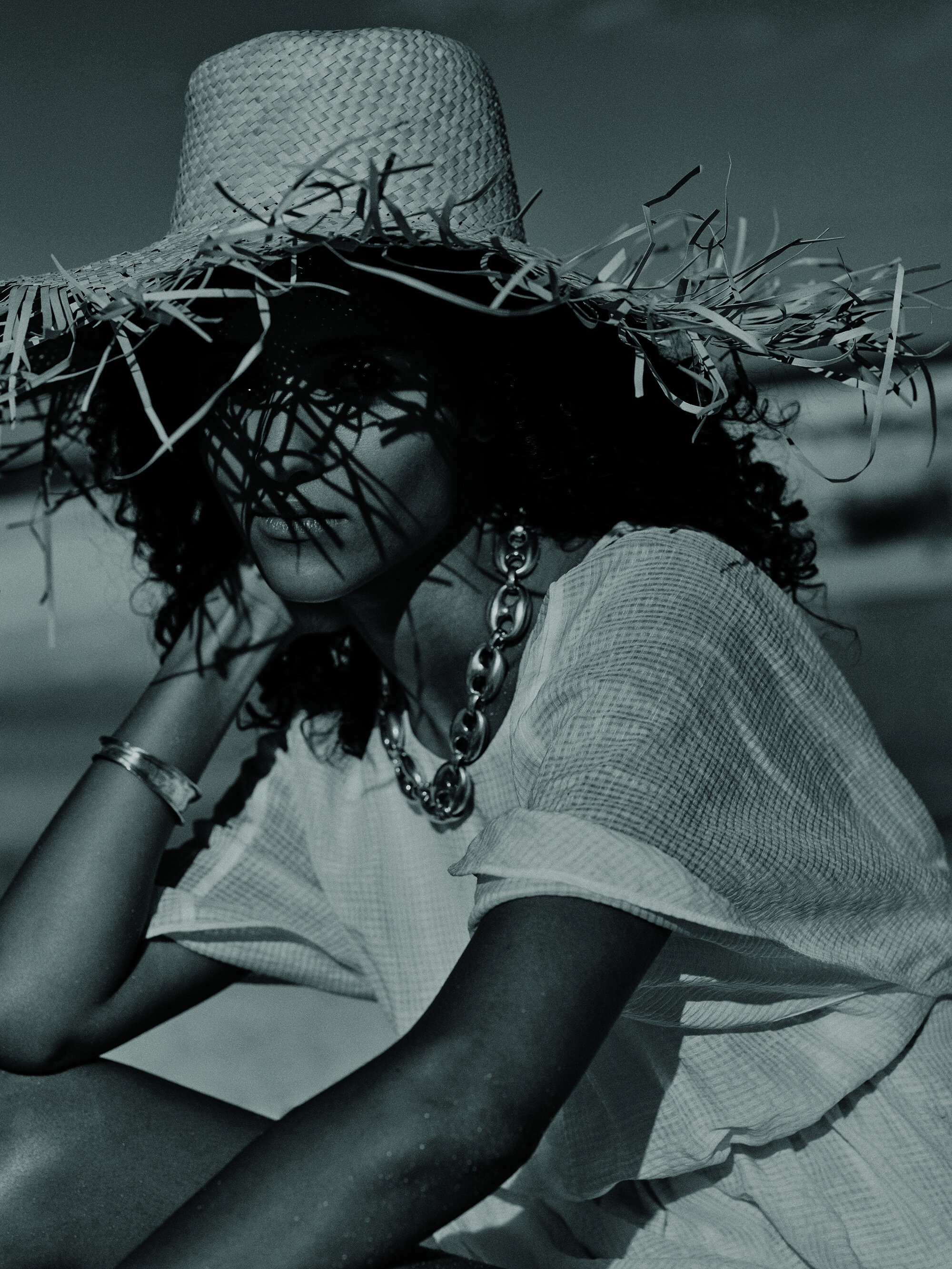 Woman poses on a sandy beach wearing a large straw hat, statement necklace and casual clothing in a fashion photography style shot.