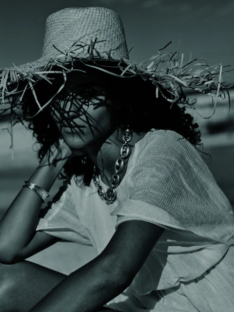 Woman poses on a sandy beach wearing a large straw hat, statement necklace and casual clothing in a fashion photography style shot.