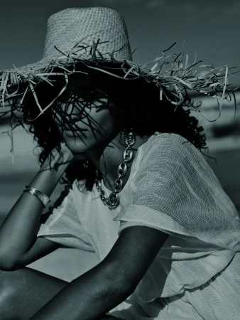 Woman poses on a sandy beach wearing a large straw hat, statement necklace and casual clothing in a fashion photography style shot.