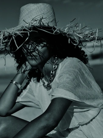 A woman with curly hair sitting on a beach, wearing a floppy straw hat, sheer white dress, and chunky jewelry in a fashion editorial.