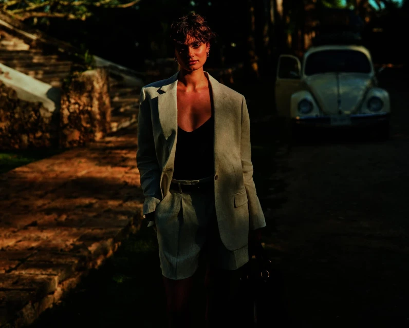 Woman poses in linen suit with vintage car backdrop on sun-drenched path. Dark hair and natural light enhance this outdoor fashion shot.