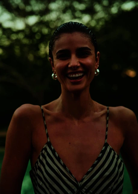 Portrait of a smiling woman in a striped dress, outdoors. Close-up shot with blurred green background of trees. She is wearing hoop earrings.