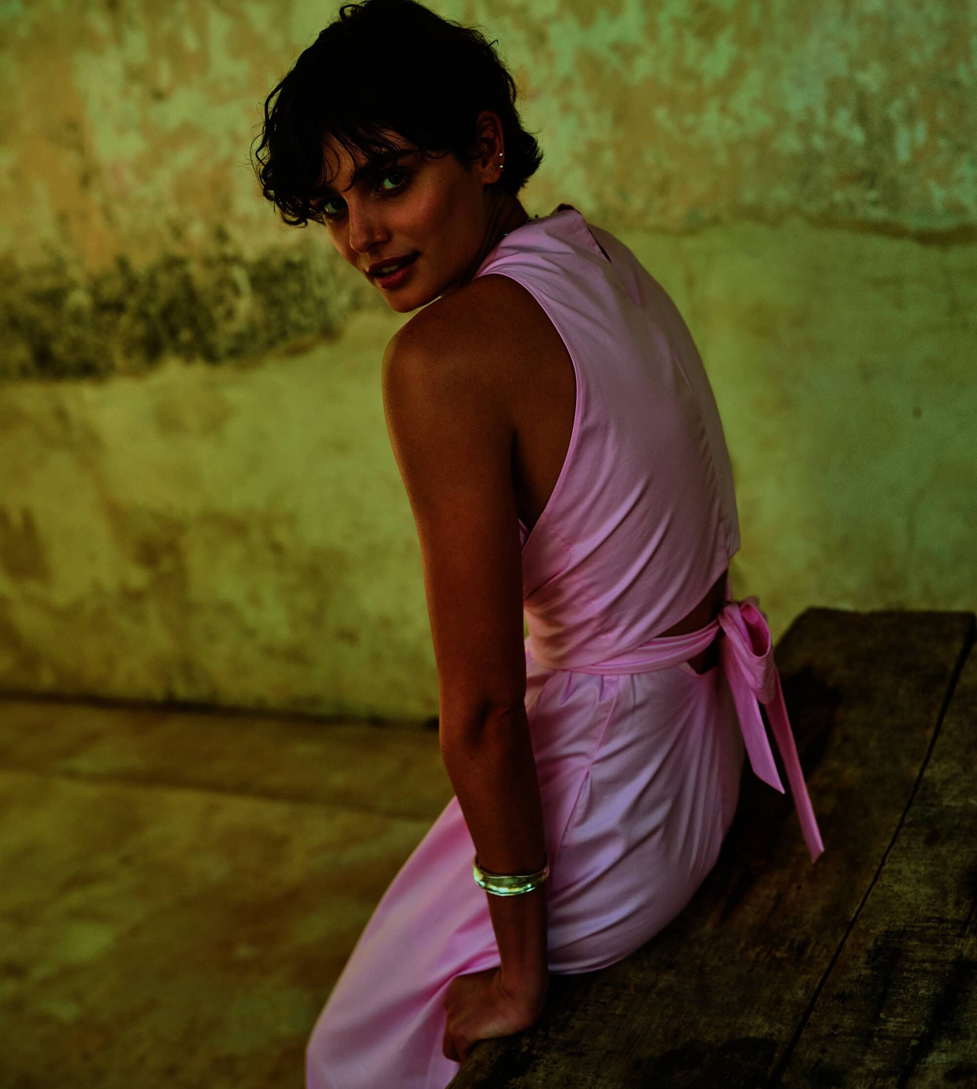 Brunette adult female fashion model sits indoors on a wooden surface, glancing backward at the camera. She wears a light pink outfit.