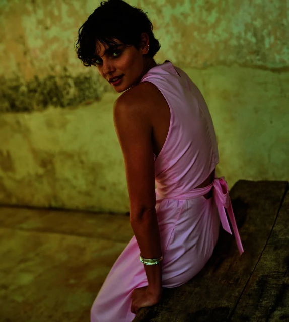 Brunette adult female fashion model sits indoors on a wooden surface, glancing backward at the camera. She wears a light pink outfit.
