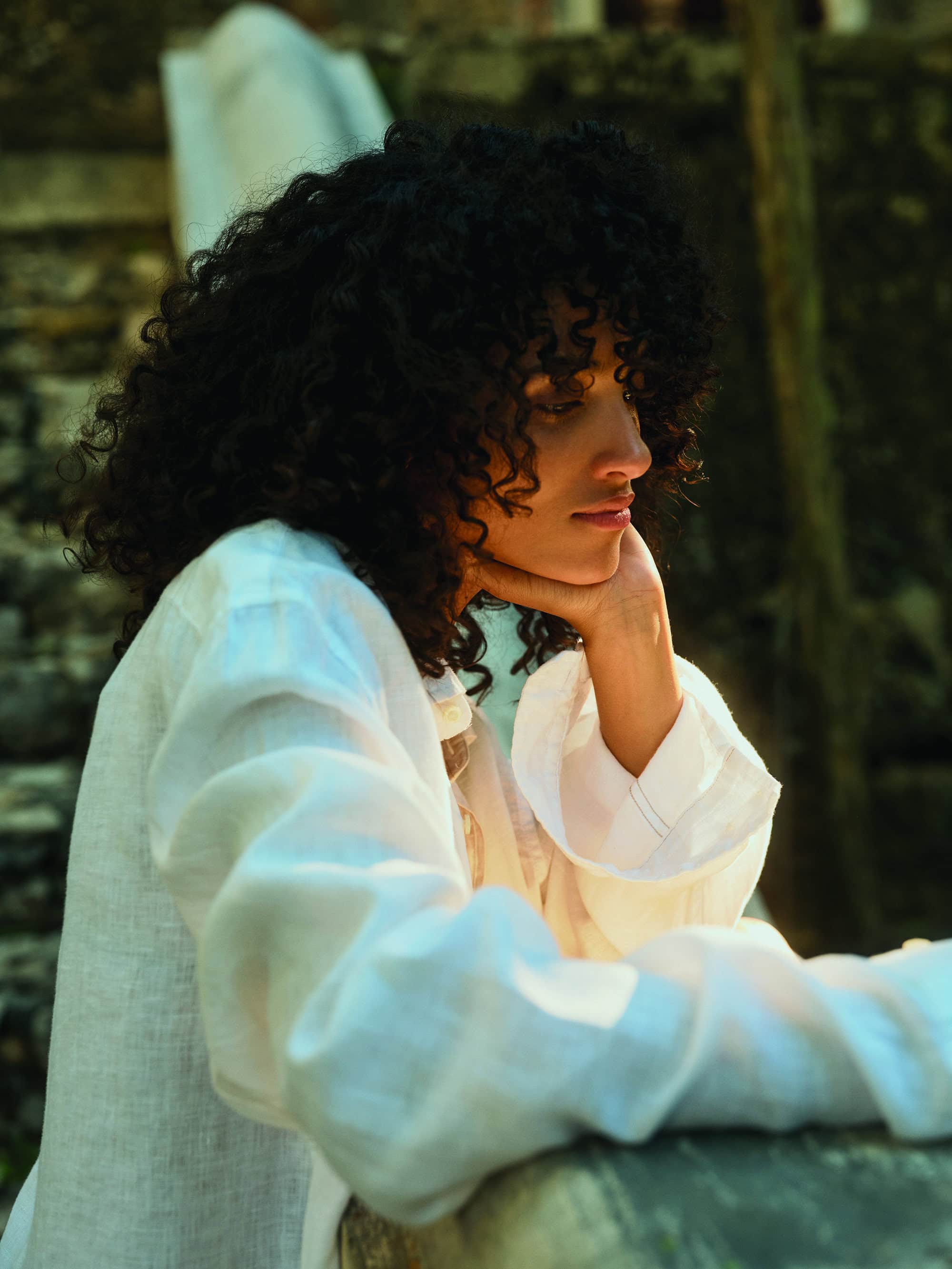 Portrait of a young woman with dark curly hair in a white long-sleeved shirt leaning on her hand outdoors on a sunny day.
