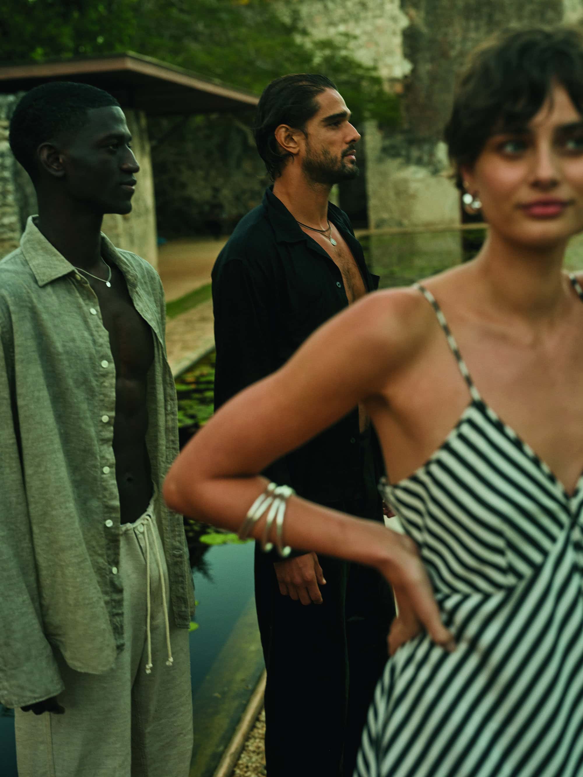Three adults in casual linen attire pose outdoors near water. One Black male in sage shirt and white pants, woman in striped dress.