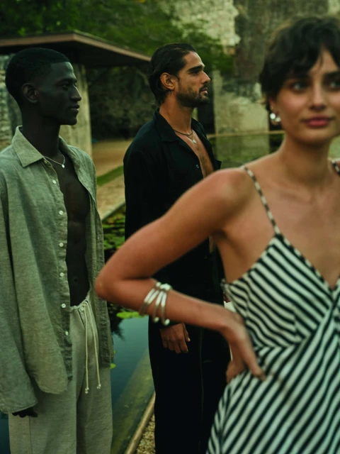 Three adults in casual linen attire pose outdoors near water. One Black male in sage shirt and white pants, woman in striped dress.