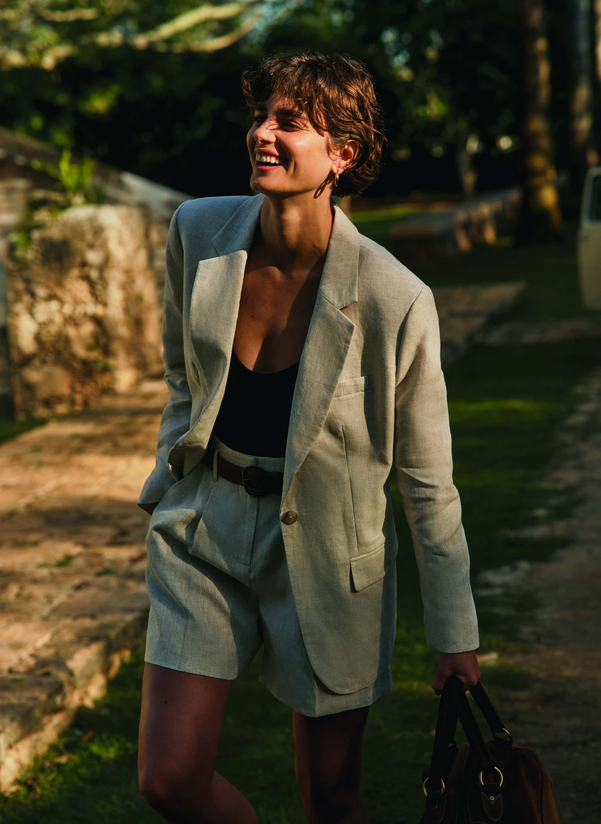 Adult woman in shorts suit smiles, walking in a garden carrying a bag, with natural light and lush green foliage background.