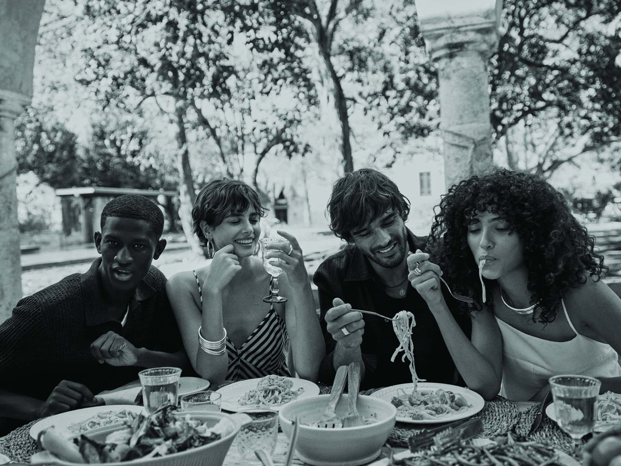Four young adults share a black and white outdoor meal at a table with salad, pasta, and drinks, set against a blurred natural backdrop.
