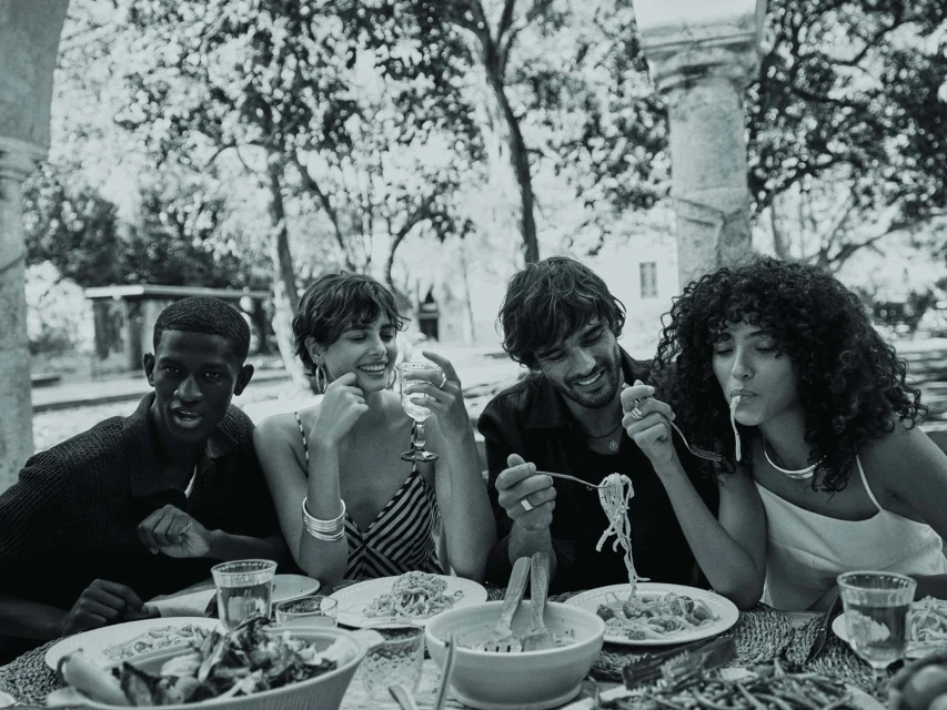 Four young adults share a black and white outdoor meal at a table with salad, pasta, and drinks, set against a blurred natural backdrop.