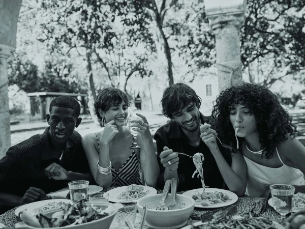 Four young adults share a black and white outdoor meal at a table with salad, pasta, and drinks, set against a blurred natural backdrop.