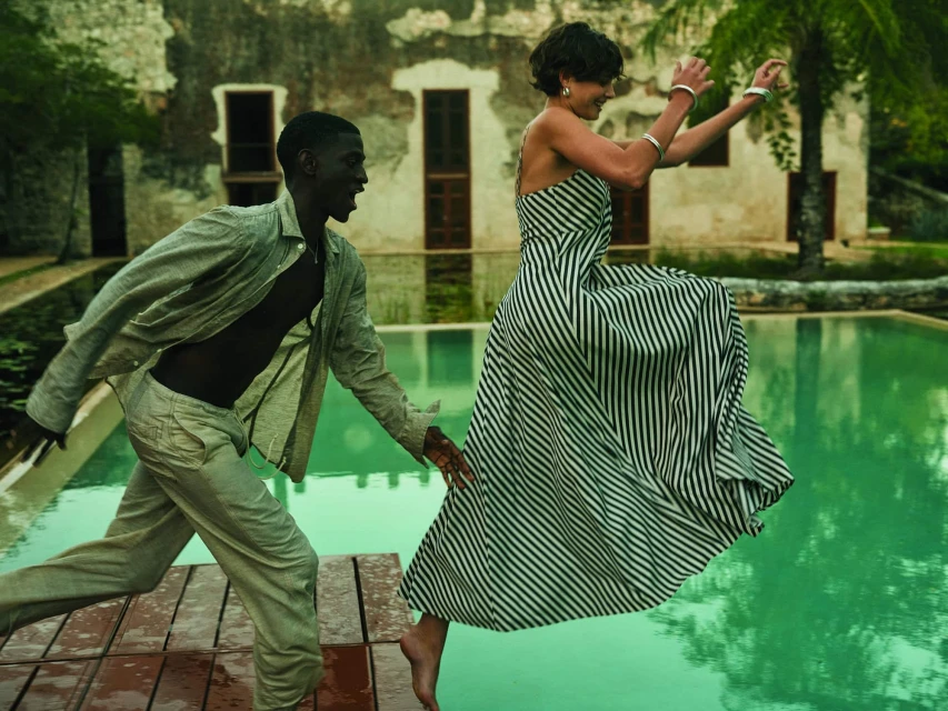 Woman in striped dress leaping over a turquoise pool toward a man on a wooden deck, with a rustic building and trees in the background.