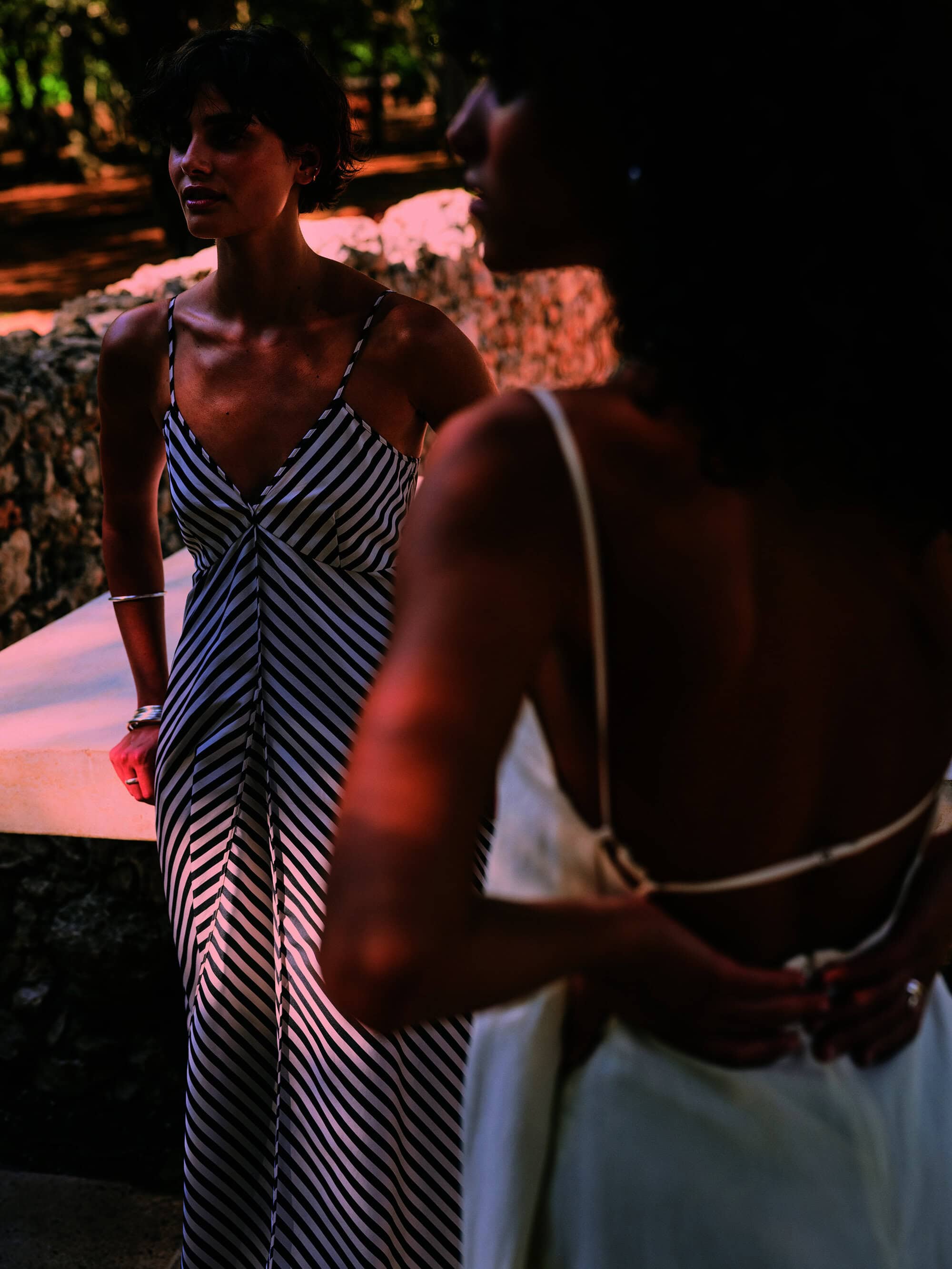 Two women in summer dresses stand outside in filtered sunlight. One dress features bold black and white stripes, jewelry visible.