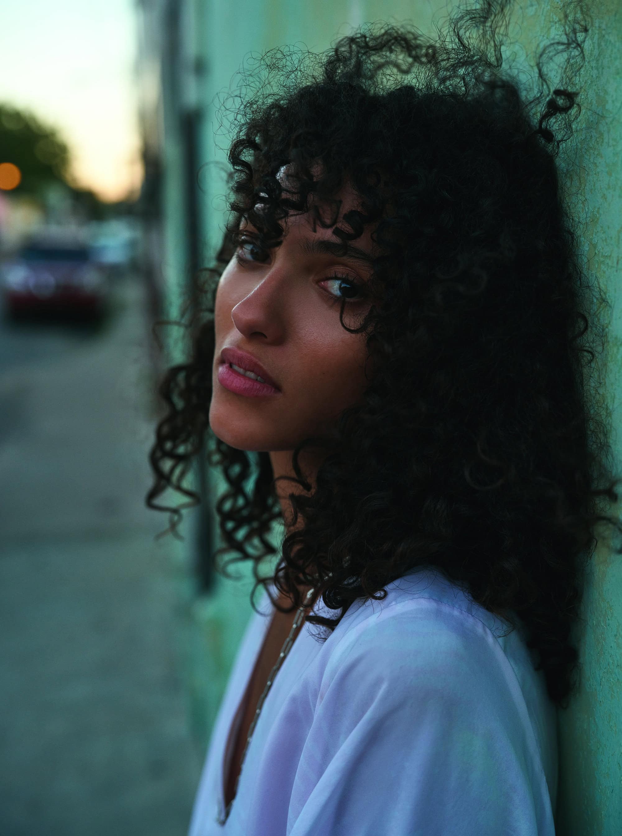 Portrait of a beautiful young adult female with curly dark hair, looking up with a white shirt against an aqua-colored wall outdoors.