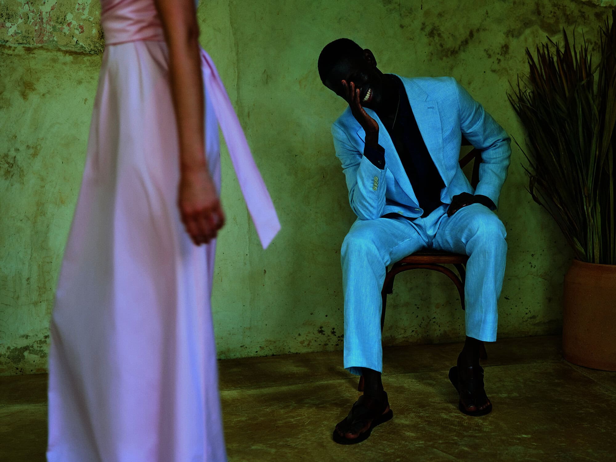 A dark-skinned man in a blue suit sits and smiles with a blurred woman in pink dress in the background and green wall. Fashion.