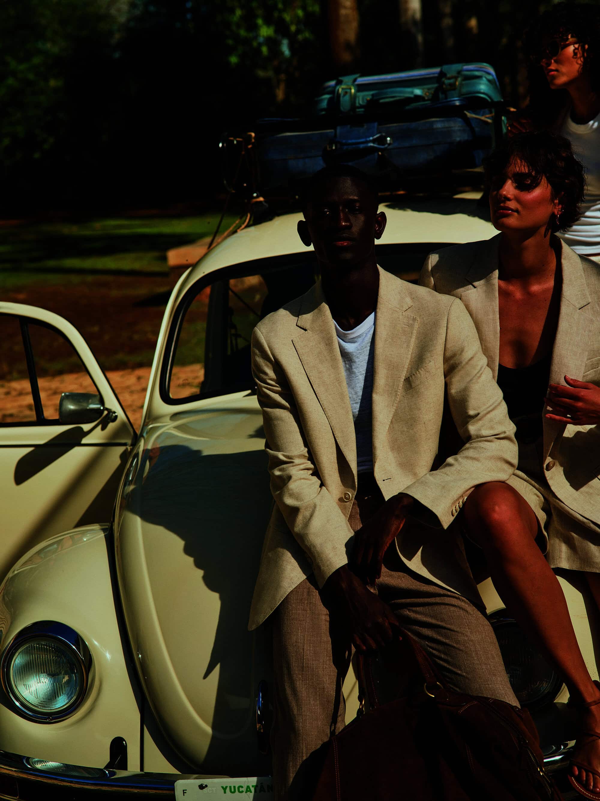 Group of three people with a vintage Volkswagen Beetle packed with luggage, posing outdoors in matching tan blazers and trousers.