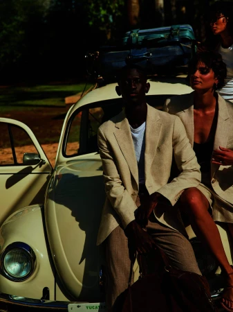 Group of three people with a vintage Volkswagen Beetle packed with luggage, posing outdoors in matching tan blazers and trousers.