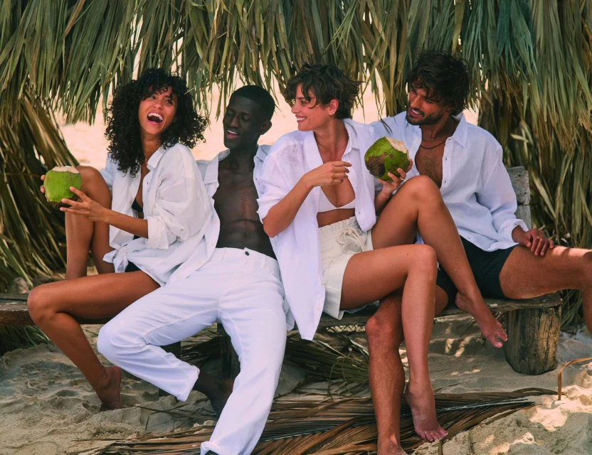 Four people in white shirts pose on a sandy beach holding coconuts under a thatched roof in bright daylight, in a medium, eye-level shot.