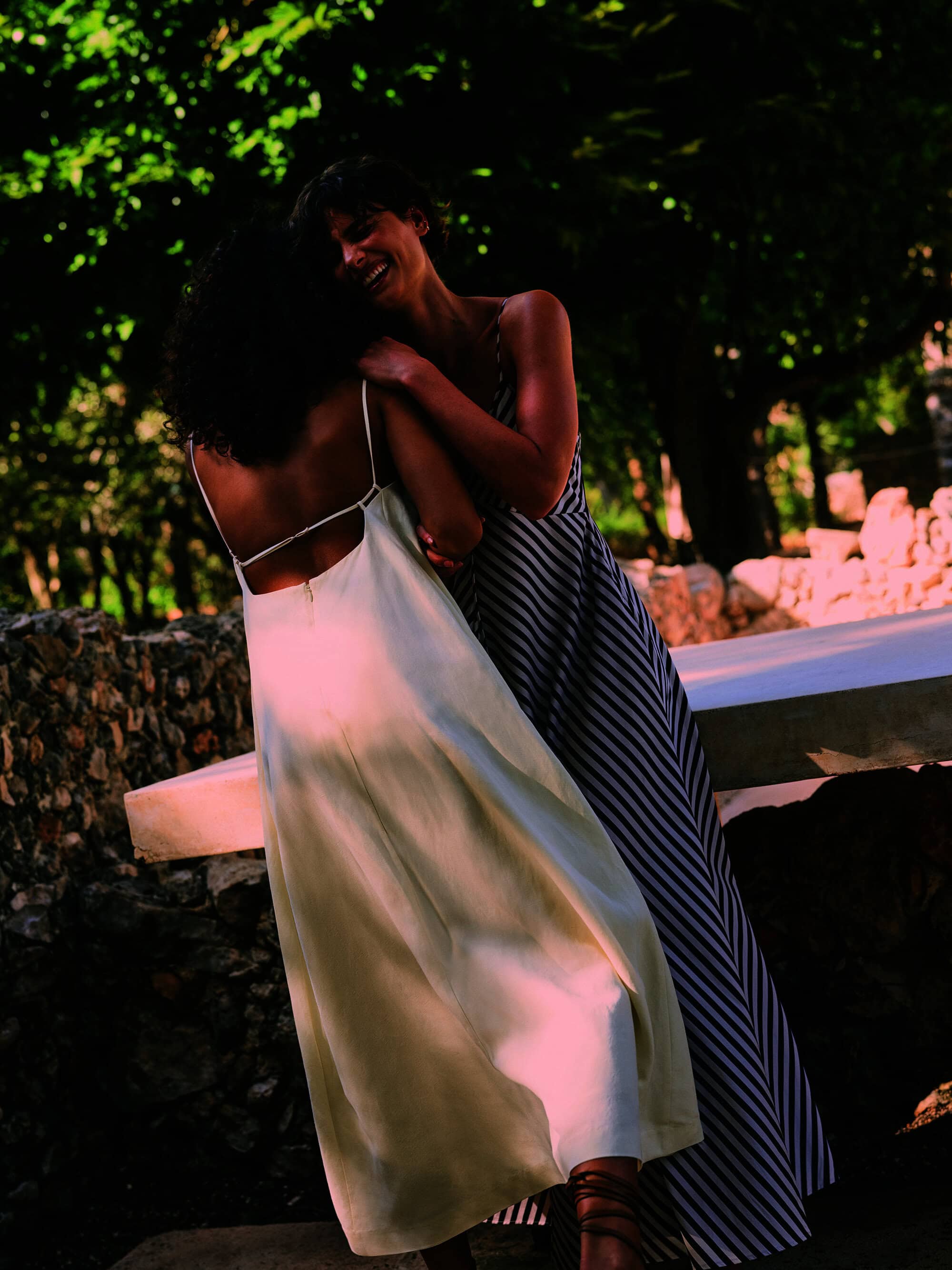Two women in dresses embrace outdoors in natural light. One woman faces away, showing the back of her dress; the other smiles.