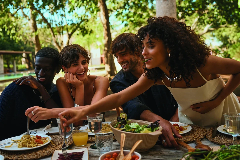 Four smiling adults enjoying an outdoor meal at a wooden table laden with pasta, salad, juice, and tomatoes. Natural light, leafy trees.