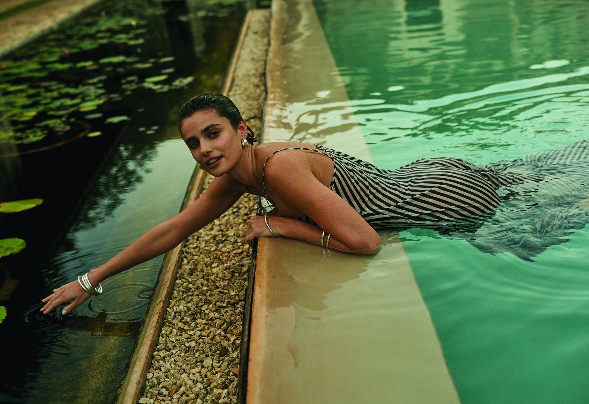 A woman in a striped dress is partially submerged in a pool, gently touching the water near lily pads. She gazes towards the camera.