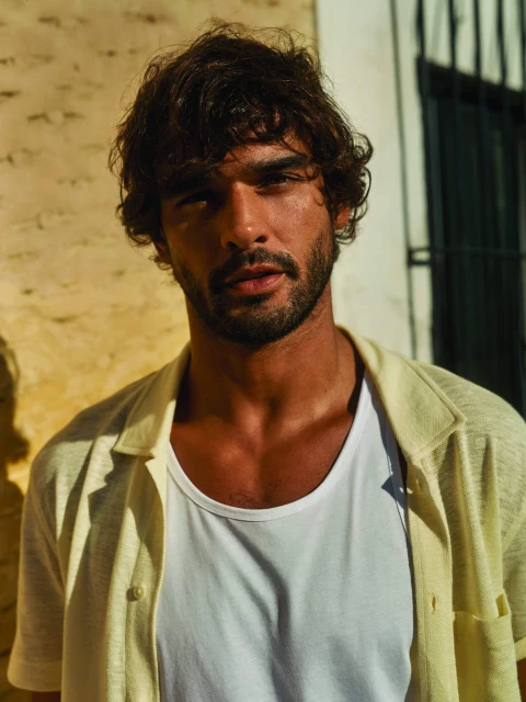 Portrait of a dark-haired man with a beard, wearing a white t-shirt and an open yellow shirt, against a light yellow wall.