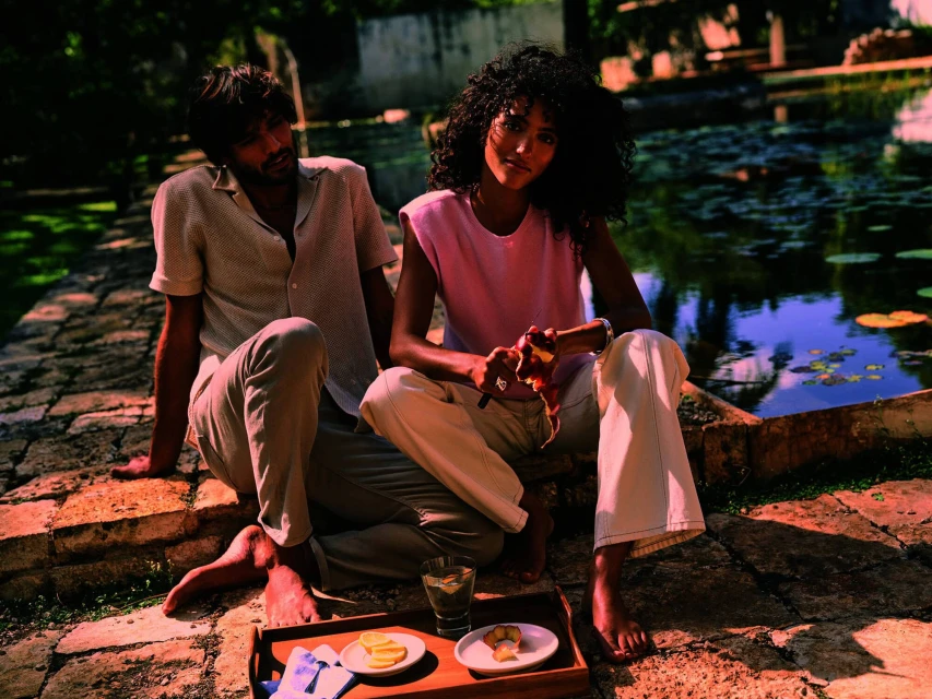 A man and a woman sit together outdoors in natural light, enjoying fruit and drinks near a pond with trees, showcasing a relaxed lifestyle.