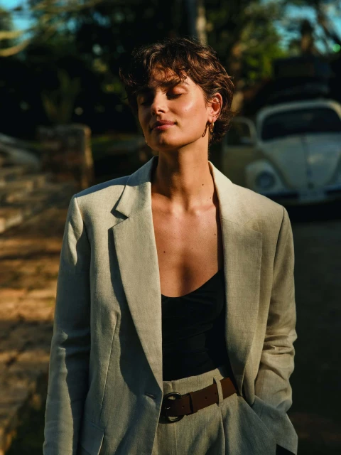 A woman in a blazer stands outdoors in a portrait shot with a vintage car in the background, lit by soft, natural light.