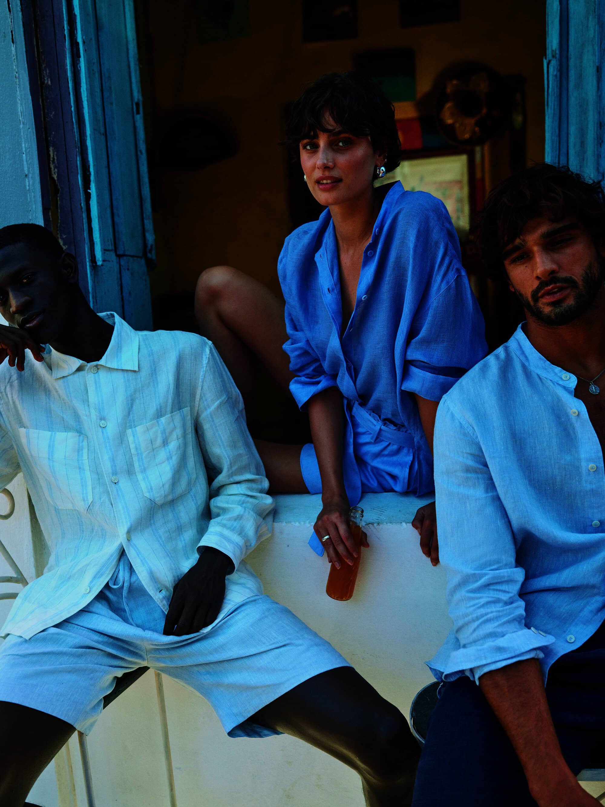 Three people in blue and white casual linen shirts and shorts are gathered indoors. The background features a partially-visible blue wall.
