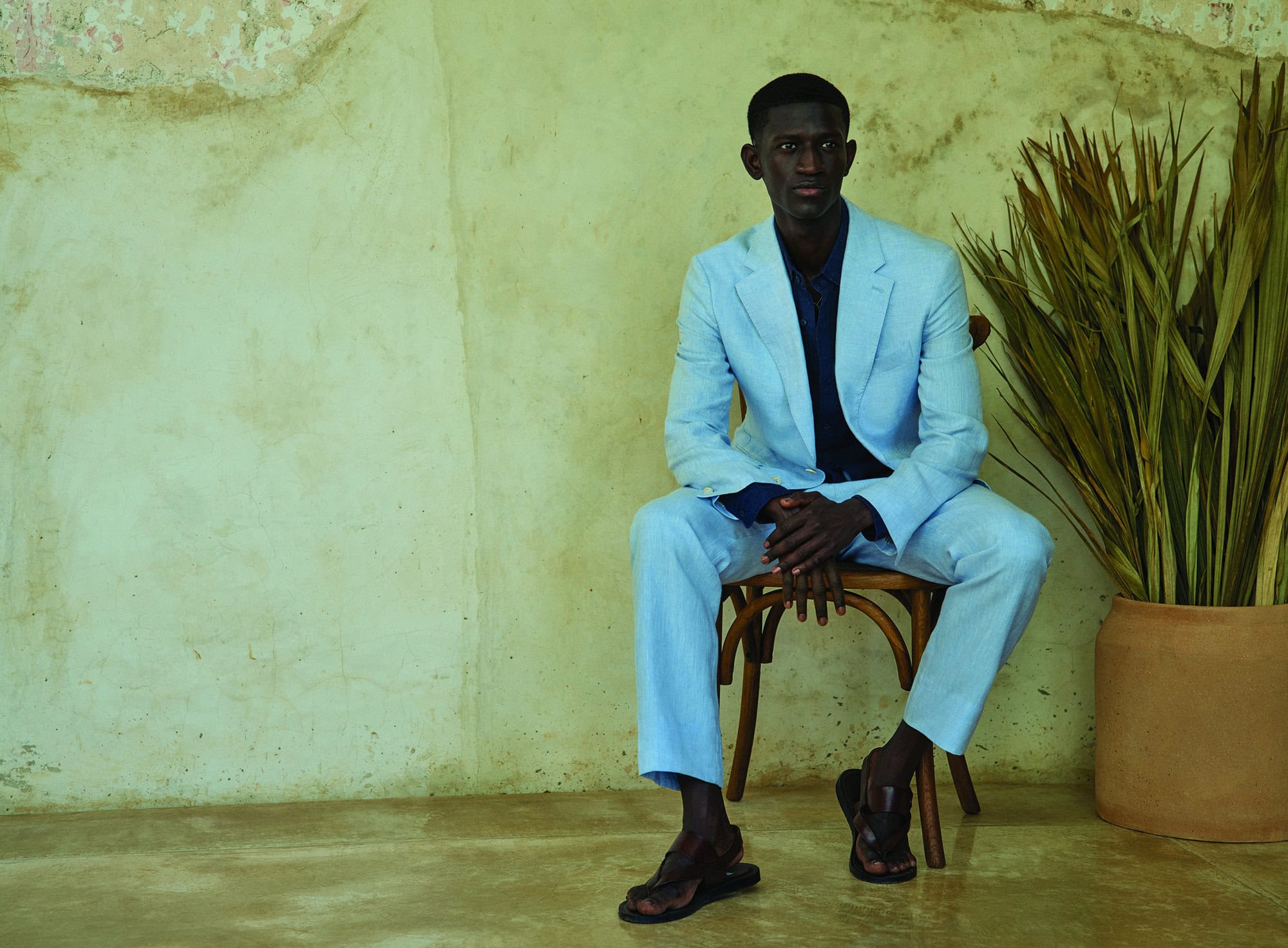 A Black male model in a light blue suit sits on a wooden chair next to a plant, in a studio with a textured wall.