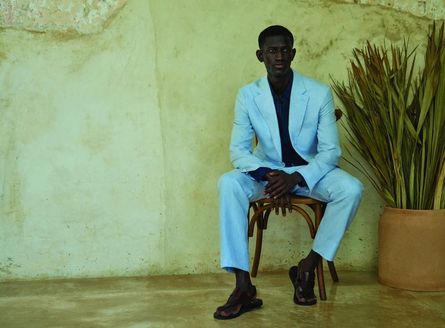 A Black male model in a light blue suit sits on a wooden chair next to a plant, in a studio with a textured wall.