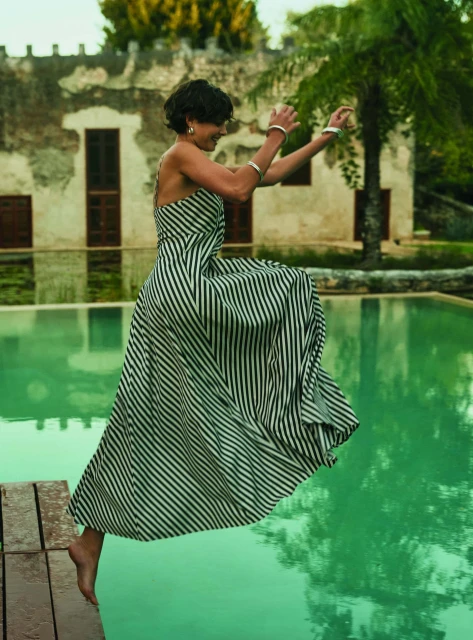 Woman in striped dress jumps near aqua pool and old building, accessorized with silver cuffs, in an outdoor, sunny setting. 