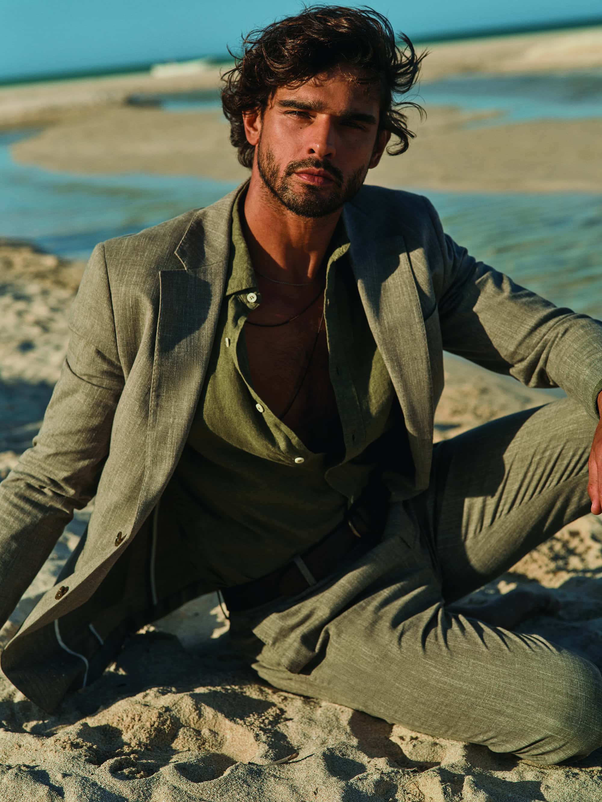 Man in a linen suit poses on a sandy beach with ocean water in the background on a sunny day, showcasing casual elegance.
