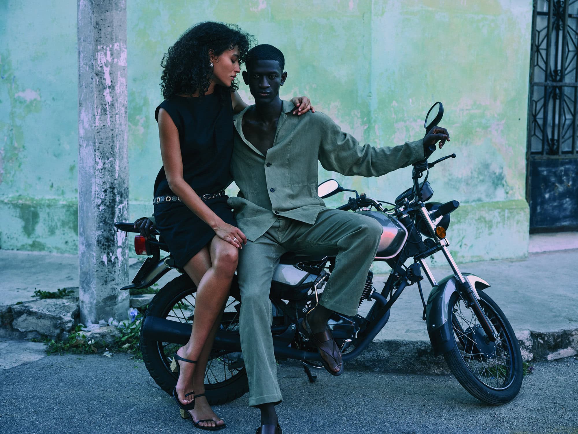 Couple posing on a motorcycle. A black woman in a black dress with a silver belt, and a black man in a green suit sit outside.