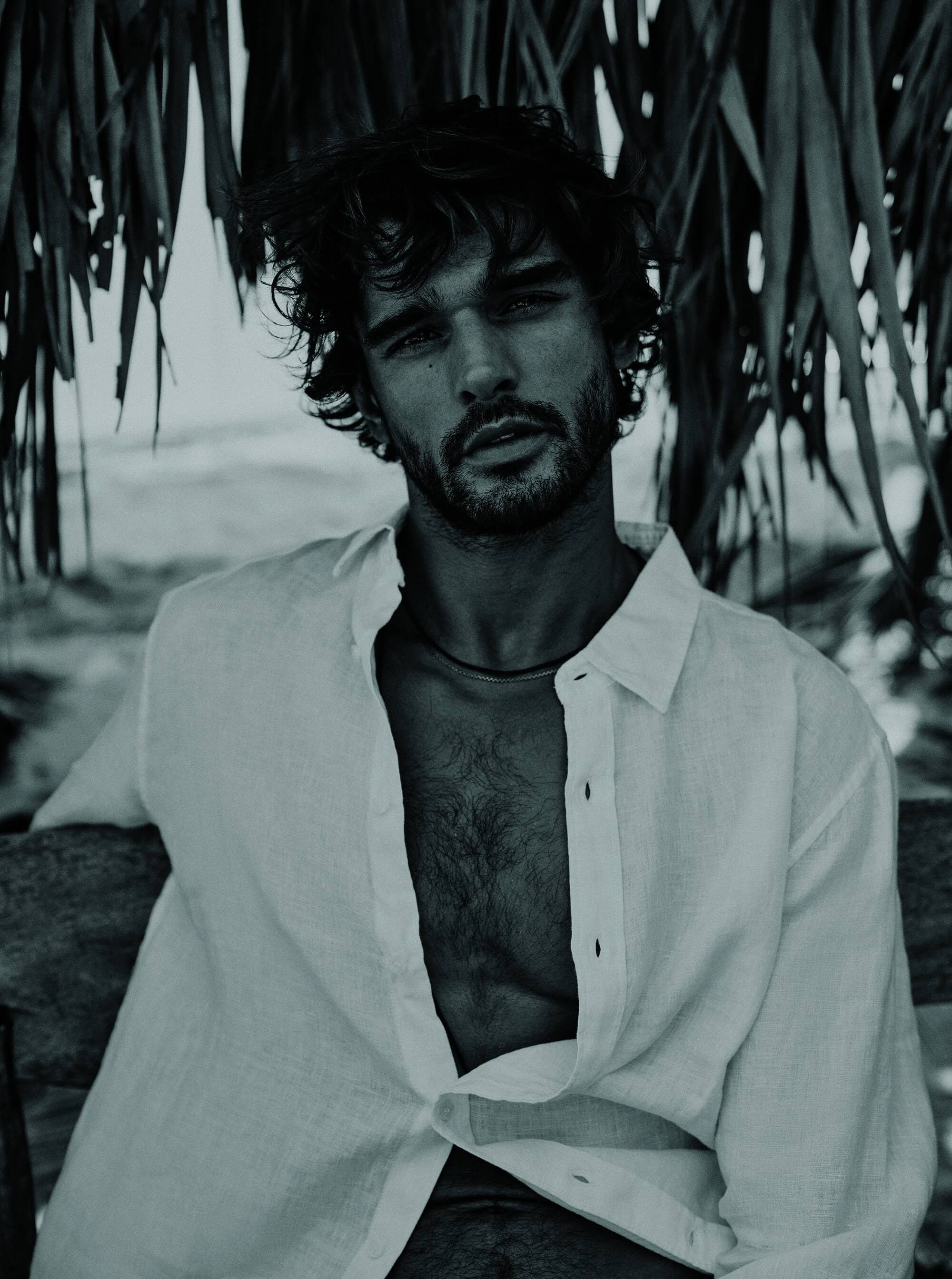 Portrait of a handsome adult male with open white shirt seated outdoors against a tropical foliage and coastal background in monochrome.