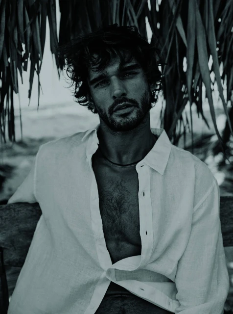 Portrait of a handsome adult male with open white shirt seated outdoors against a tropical foliage and coastal background in monochrome.