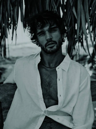 Portrait of a handsome adult male with open white shirt seated outdoors against a tropical foliage and coastal background in monochrome.