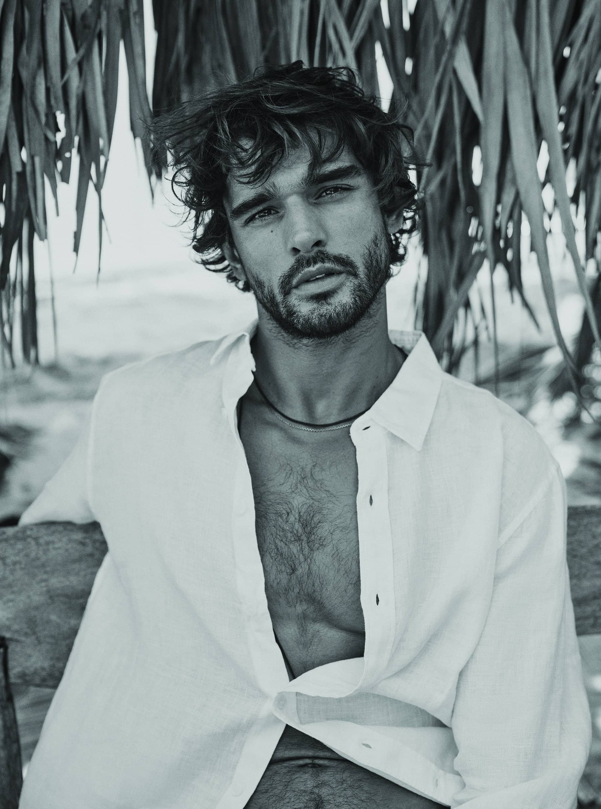 Black and white portrait of an adult male wearing a white shirt, sitting outdoors near a thatch roof and ocean background.