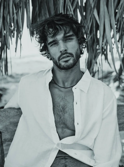 Black and white portrait of an adult male wearing a white shirt, sitting outdoors near a thatch roof and ocean background.