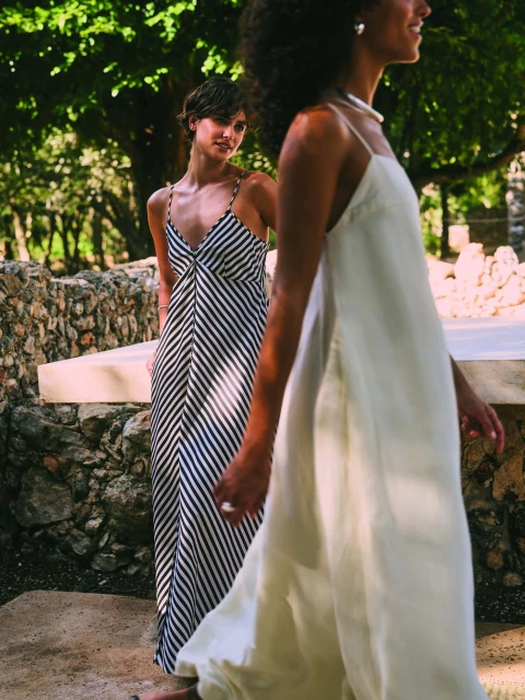 Two women in maxi dresses outdoors, one in a white dress walking, another in a striped dress looking, against a stone wall and greenery.