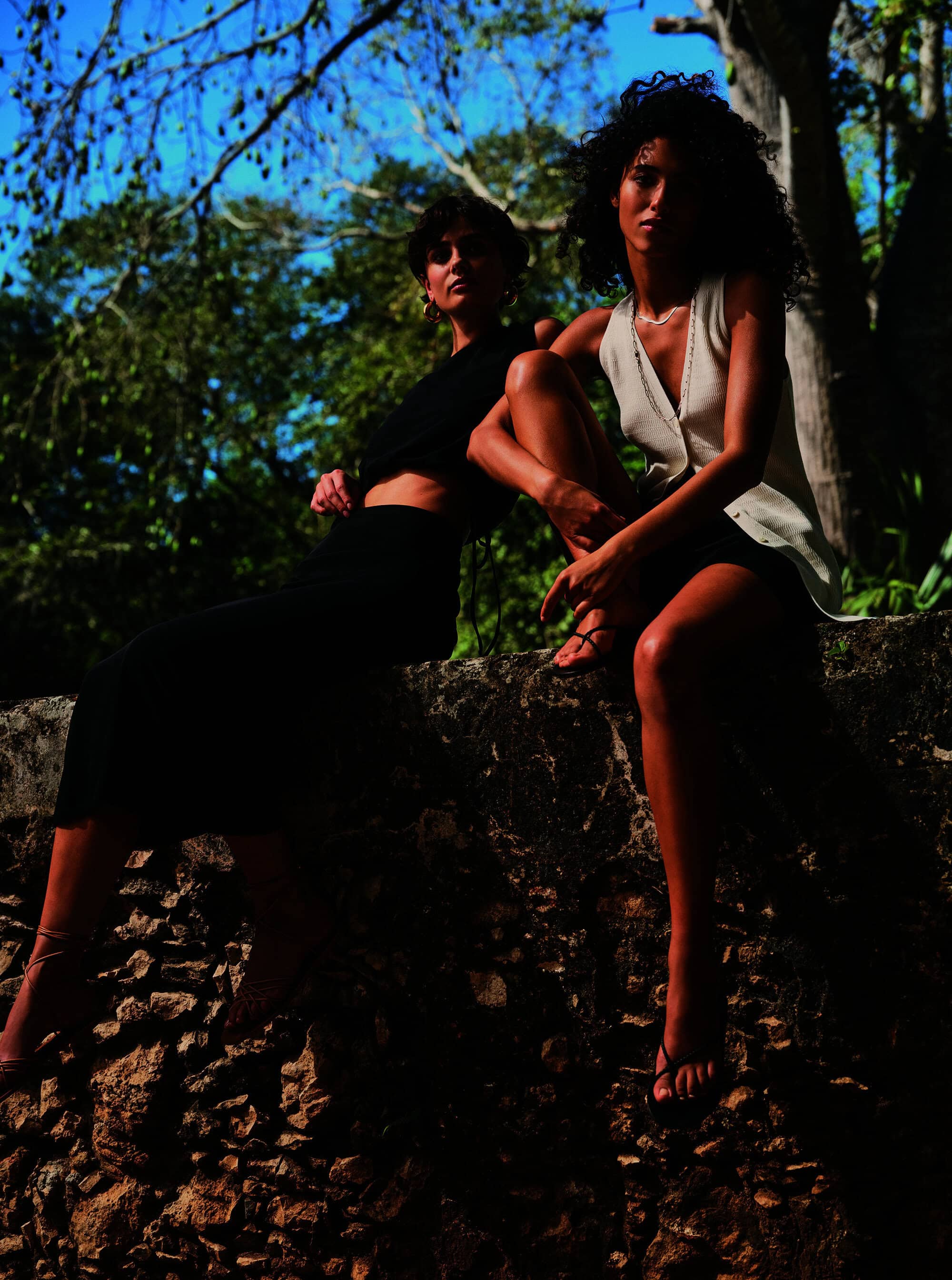 Two women sit in an outdoor setting, one in black, one in cream. They are posed on a rock wall with green trees and a blue sky backdrop.