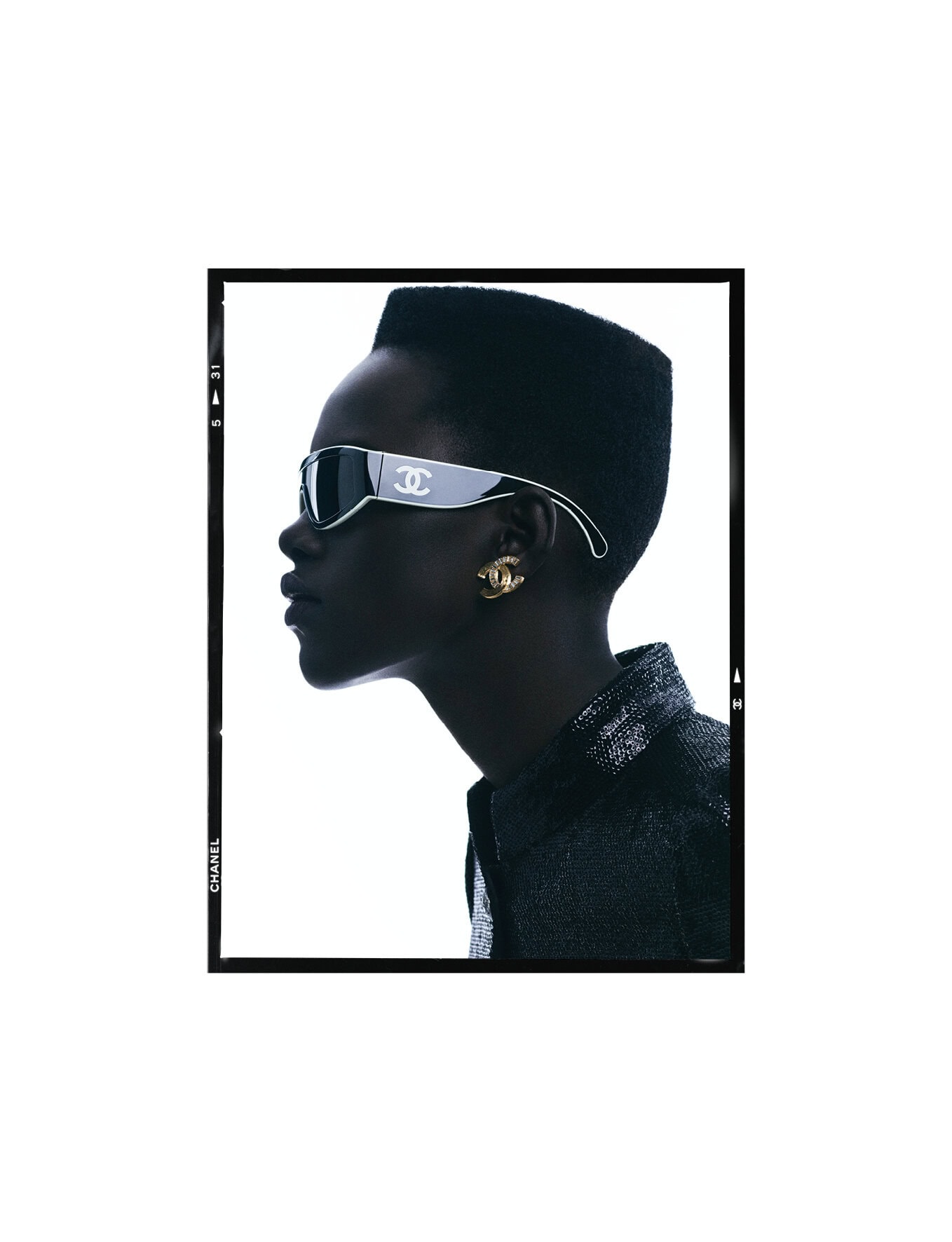 Profile shot of a stylish adult female wearing black jacket, Chanel earrings, and unique sunglasses against a white backdrop.