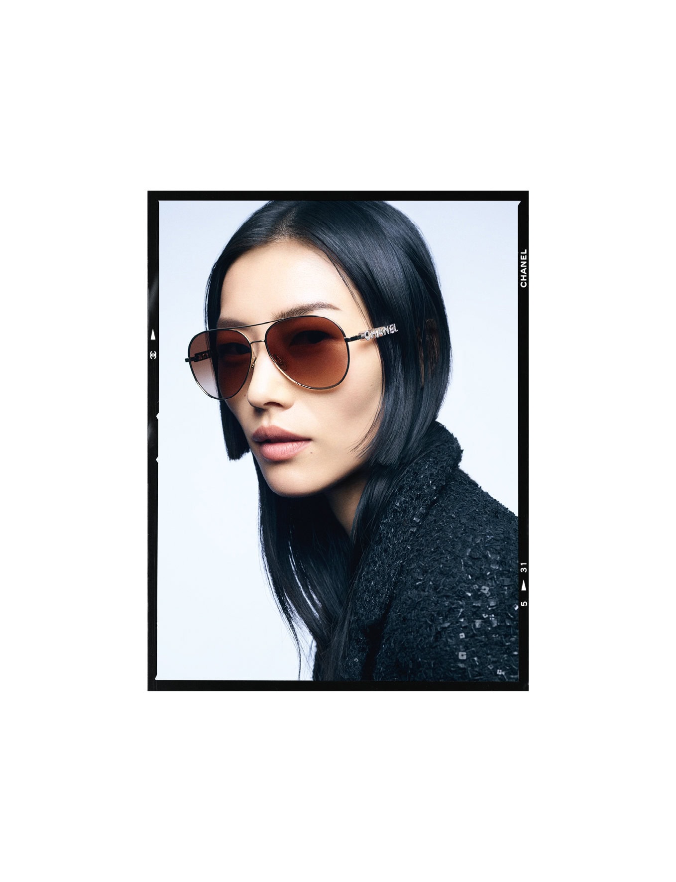 Close-up of a woman wearing aviator sunglasses and a black coat against a white background in a fashion-style studio shot.