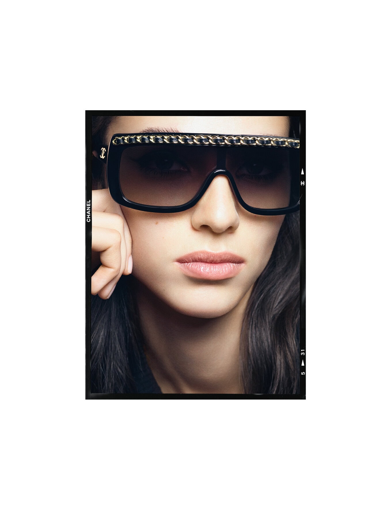 Brunette woman poses indoors wearing oversized black sunglasses with gold chain detail across the top rims in a beauty shot.