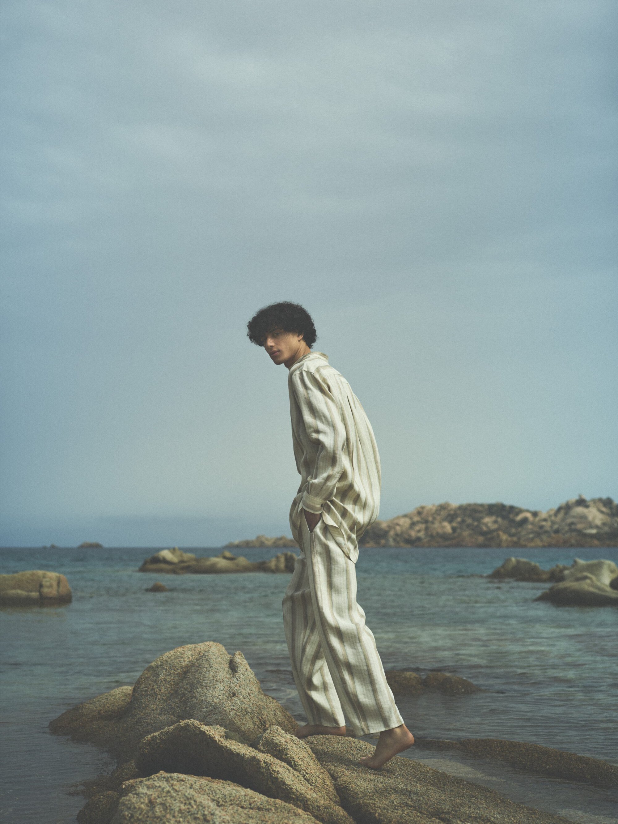 Young adult walks barefoot on rocks by the ocean wearing striped linen pants and shirt with a cloudy sky and rocky coastline.