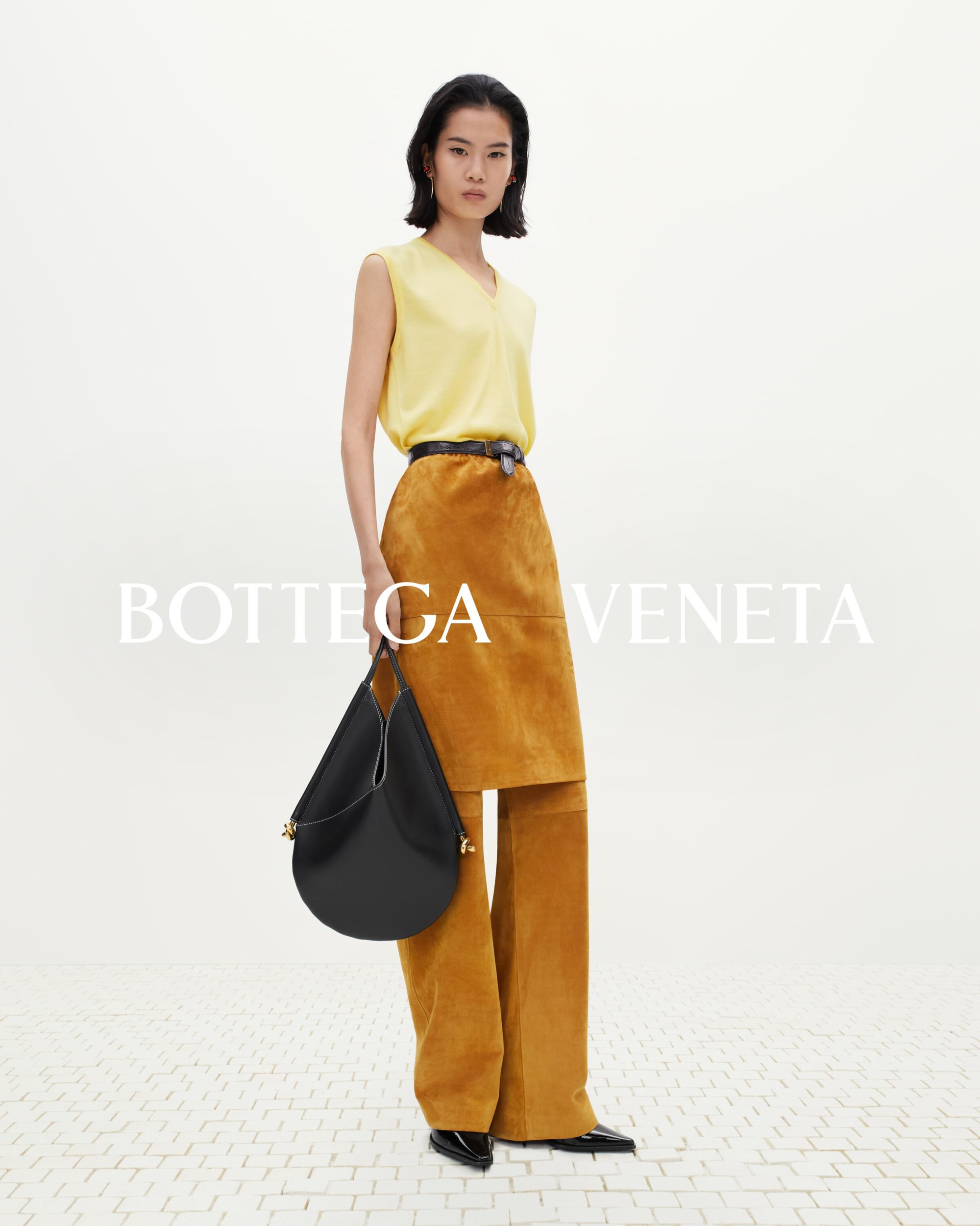 Fashion shoot featuring an adult female in a yellow top, suede skirt and pants, holding a black hobo bag against a white background.