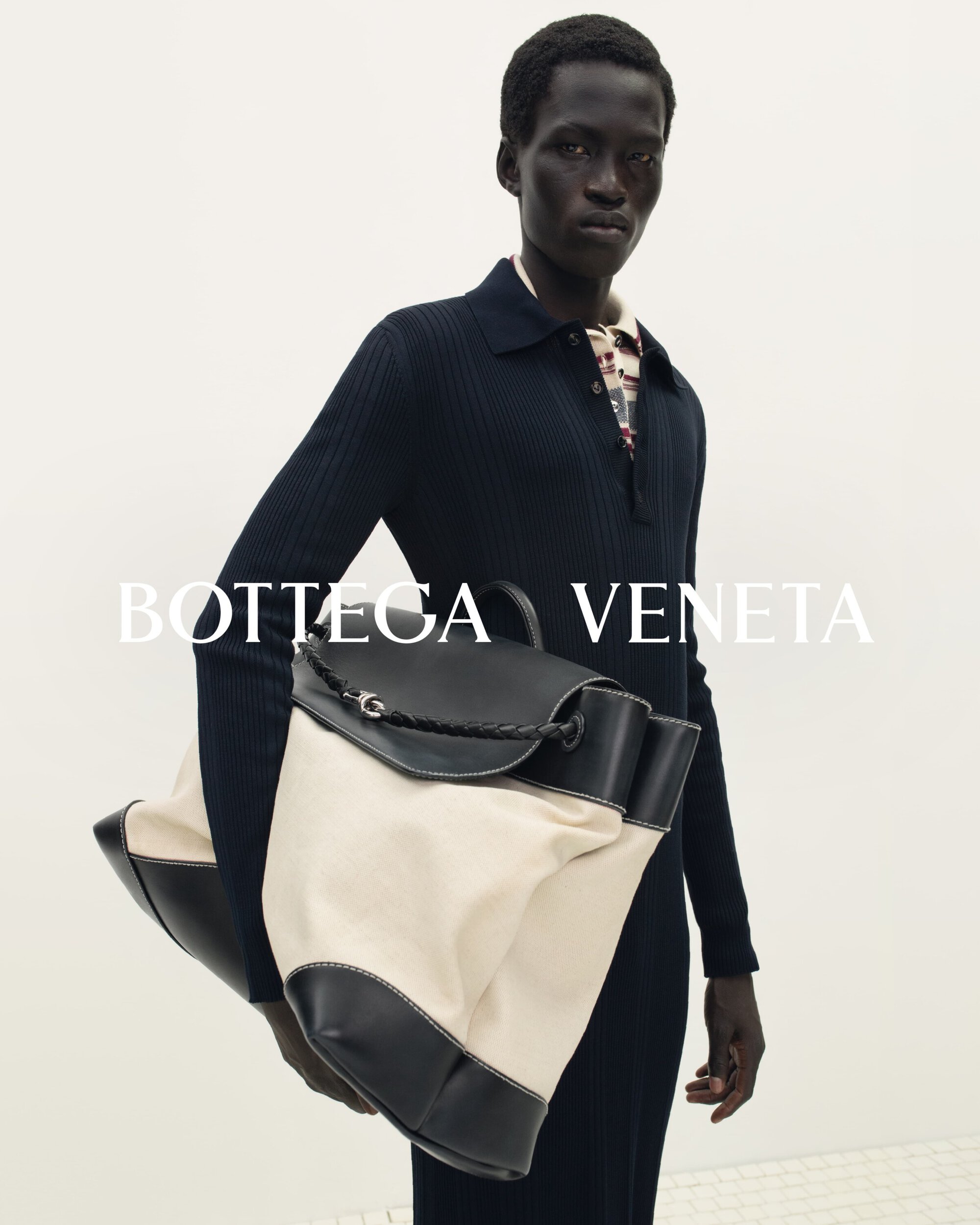 An African American male model wearing a navy sweater holds a Bottega Veneta canvas and leather bag against a white background.