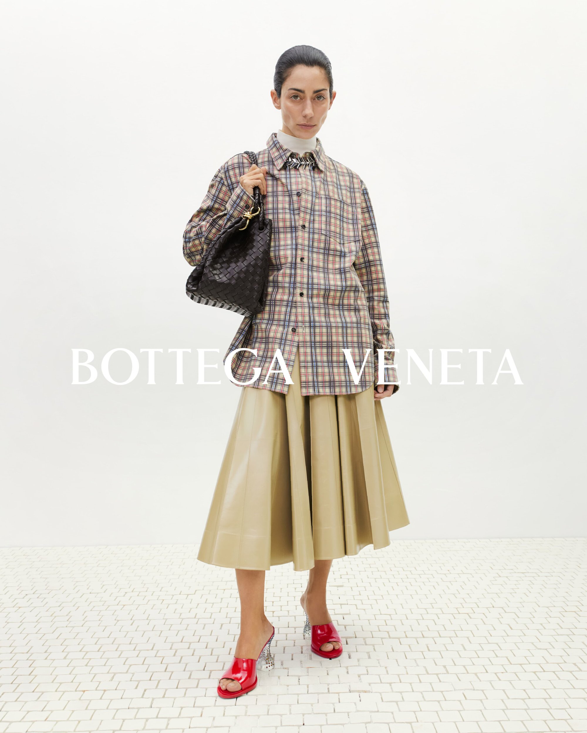 Editorial fashion shot of a female model in a plaid shirt, beige skirt, and red heels, holding a black woven handbag, against a white background.