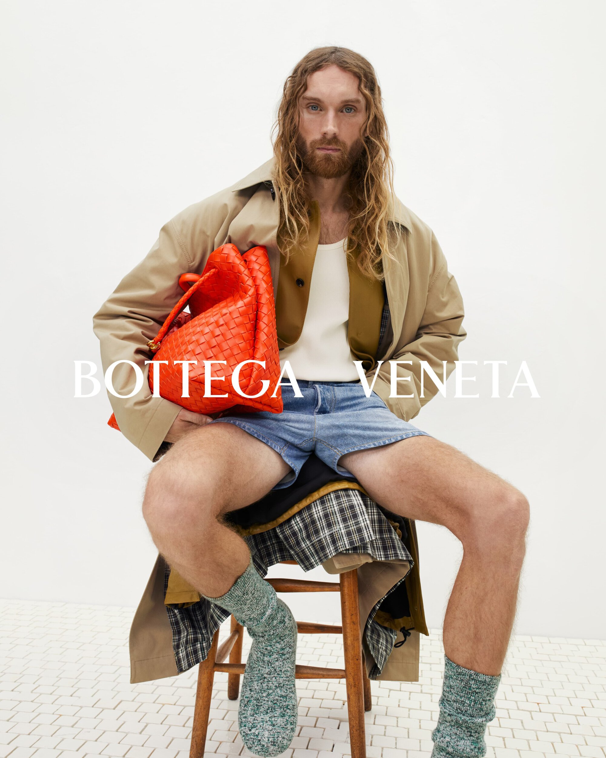Man poses with a woven orange bag in an indoor setting. He wears a beige coat, denim shorts, green socks, and sits on a wooden stool.
